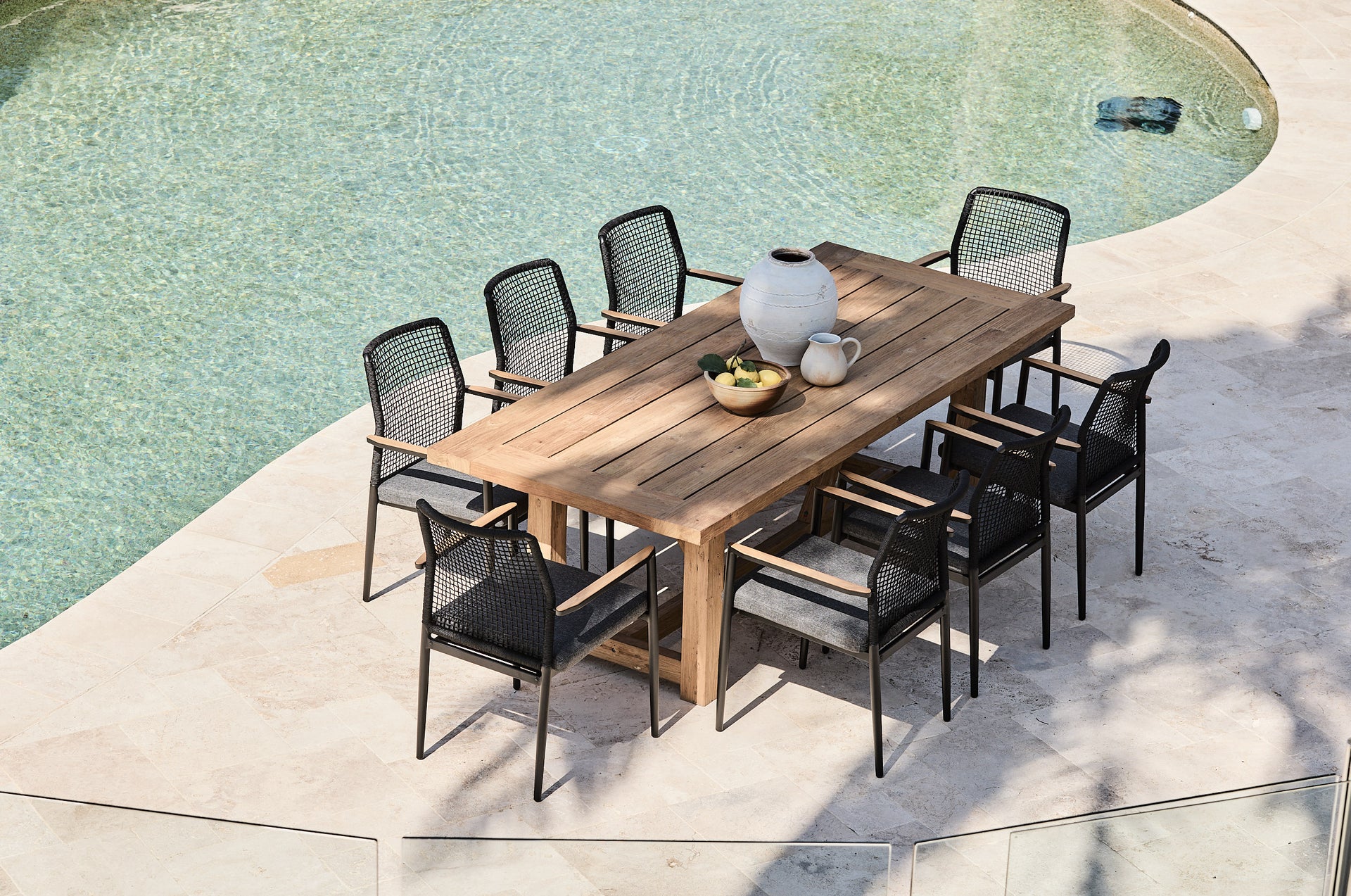 Outdoor dining set with a wooden table and black chairs by a pool.